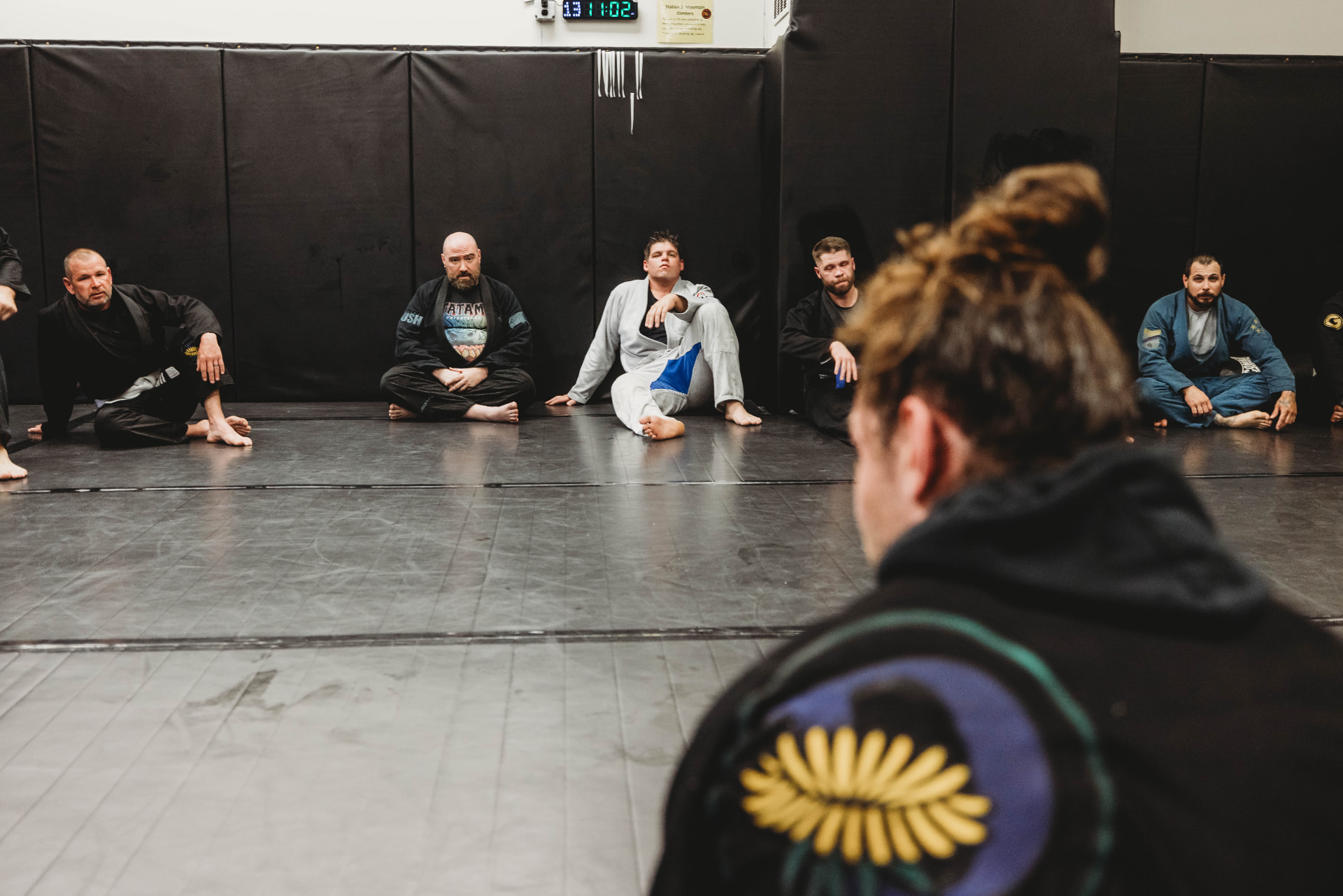 Adults sitting along the mats listening to instruction during a Brazilian Jiu-Jitsu class at Dandelion Jiu Jitsu in West Haven
