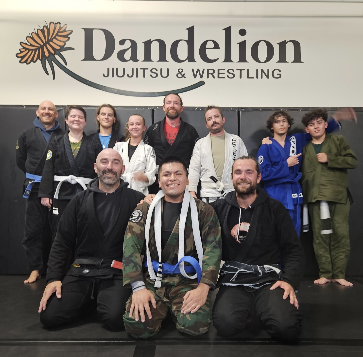Dandelion Jiu Jitsu & Wrestling students and coaches posing under the academy gym logo in West Haven